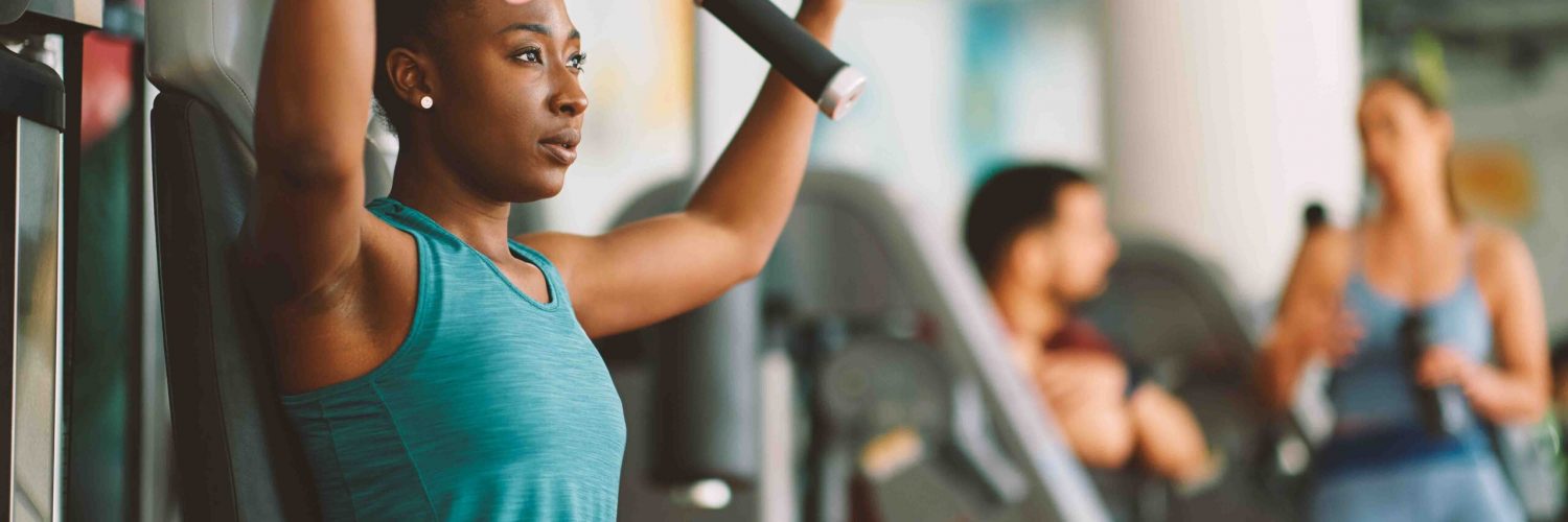 Young African American sportswoman doing chest exercises on machine while working out in gym.